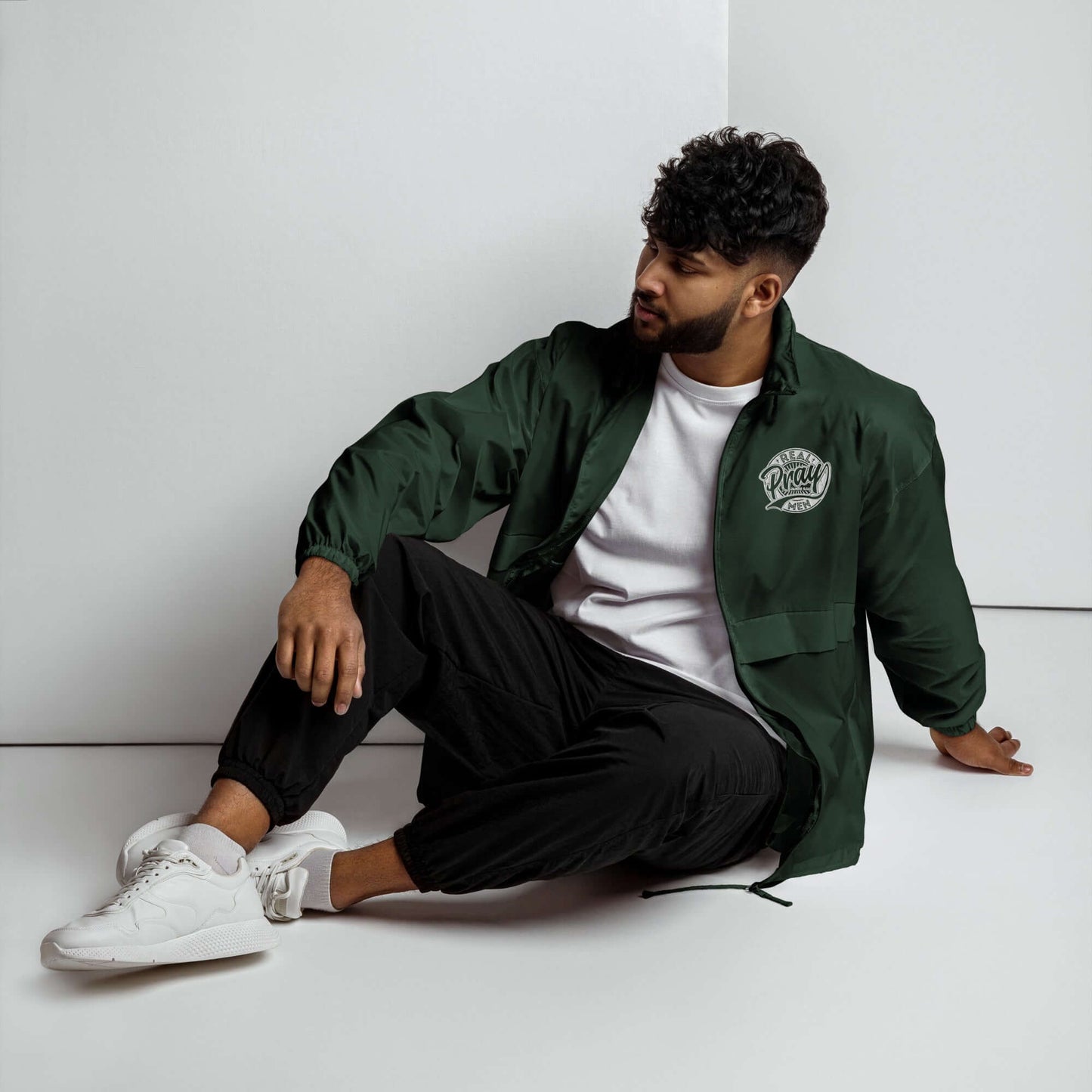 Man in green Real Men Pray windbreaker, showcasing Christian clothing style, sitting elegantly on a white backdrop.