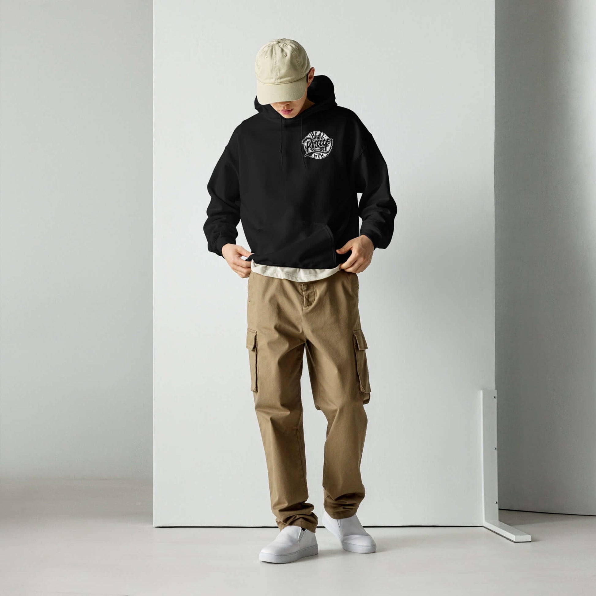 Black "Real Men Pray" hoodie worn by a young man with beige cargo pants and a cap, showcasing Christian clothing style in the UK.