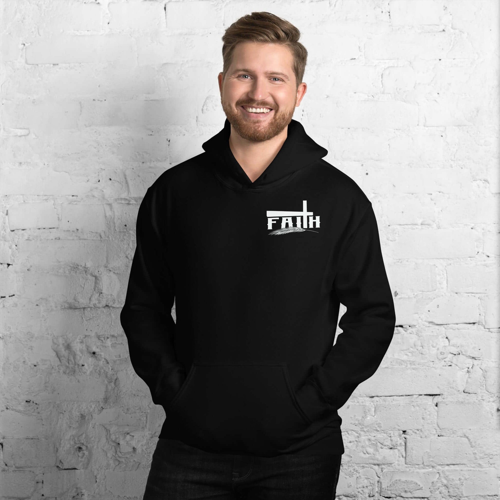 Smiling man wearing a black "Faith" hoodie with cross and feather design, inspired by Hebrews 11:1, standing against a white brick wall, Christian clothing UK.