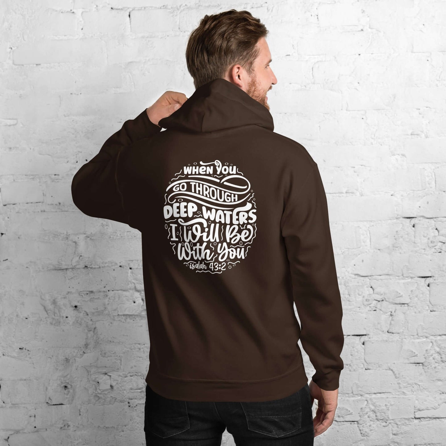 Man wearing a brown 'Through Deep Waters' hoodie with Isaiah 43:2 quote, standing against a white brick wall, Christian clothing UK.