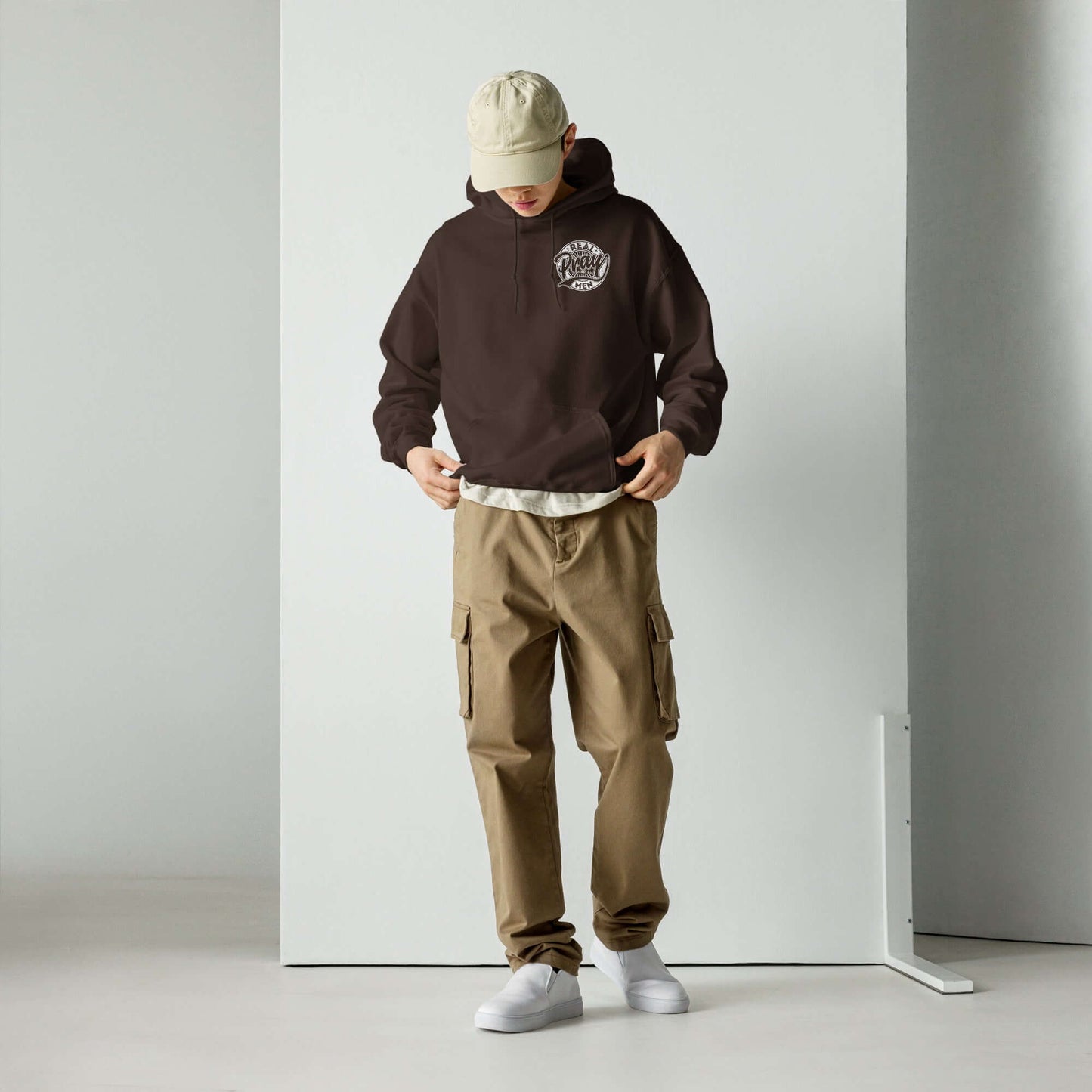 Man wearing a brown "Real Men Pray" hoodie with beige cargo pants and a cap, showcasing Christian clothing style.