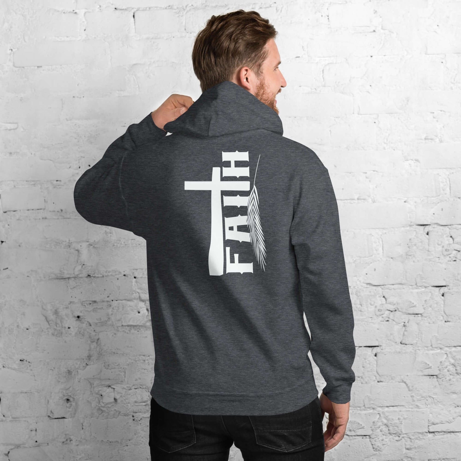 Man wearing a grey 'Faith' hoodie with a cross and feather design, inspired by Hebrews 11:1, against a white brick wall background.