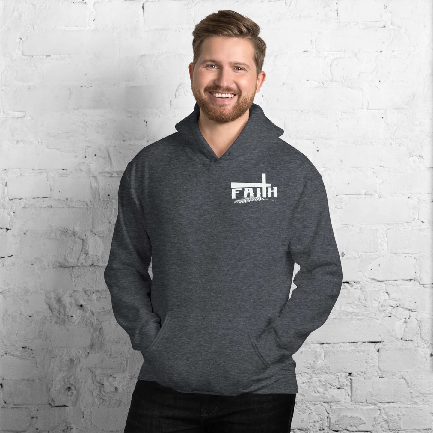 Smiling man wearing a grey Faith hoodie with cross and feather design, inspired by Hebrews 11:1, against a white brick wall, promoting Christian clothing UK.