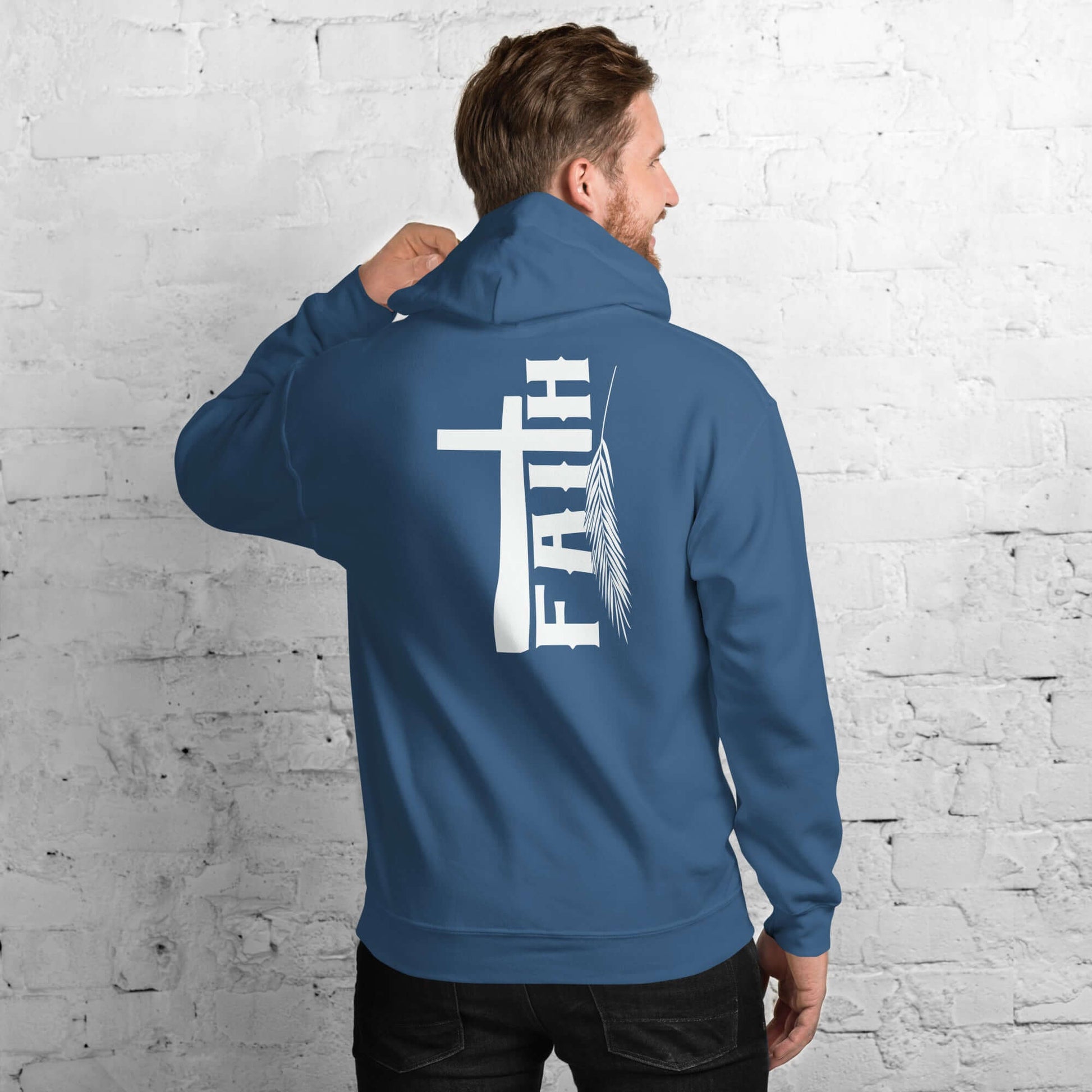 Man wearing a blue Faith hoodie with cross and feather design, inspired by Hebrews 11:1, against a white brick wall, Christian clothing UK.