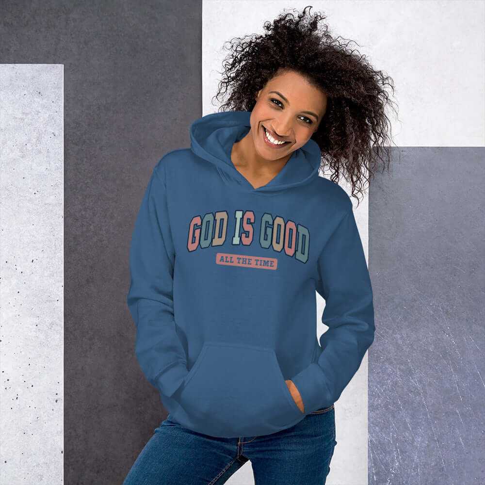 Woman smiling in a light blue "God is Good" hoodie, showcasing vibrant lettering and stylish design for Christian clothing in the UK.