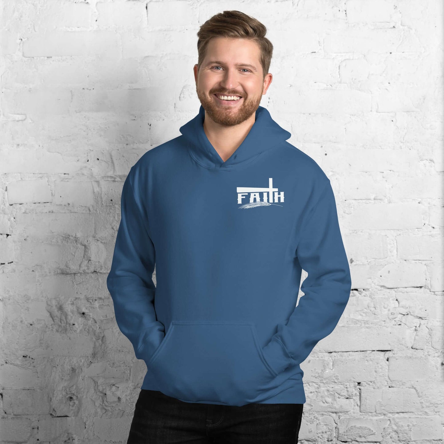 Smiling man in a blue "Faith" hoodie with cross and feather design, inspired by Hebrews 11:1, against a white brick background. Christian clothing UK.