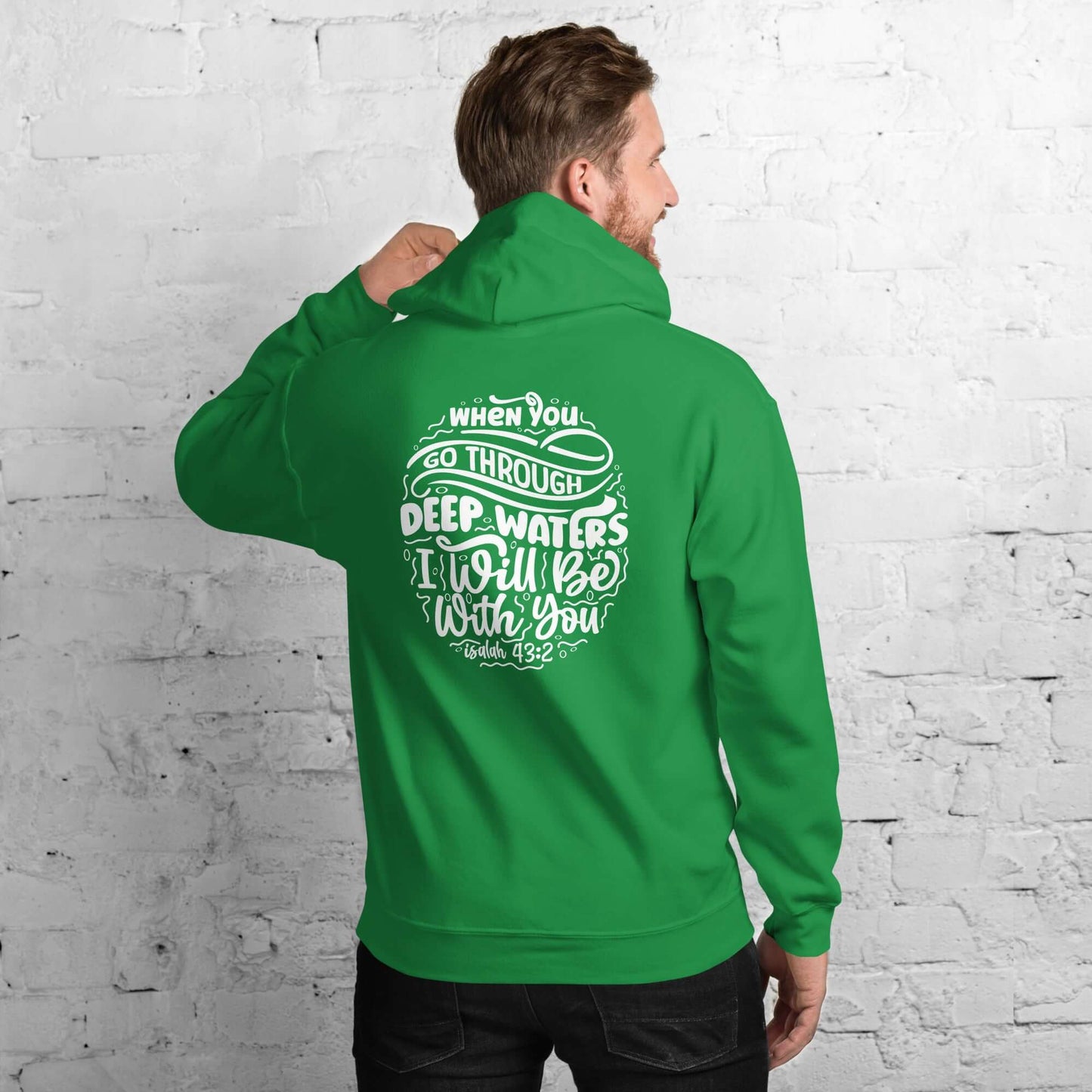 Man wearing green "Through Deep Waters" hoodie with Isaiah 43:2 quote, Christian clothing UK, standing against white brick wall.