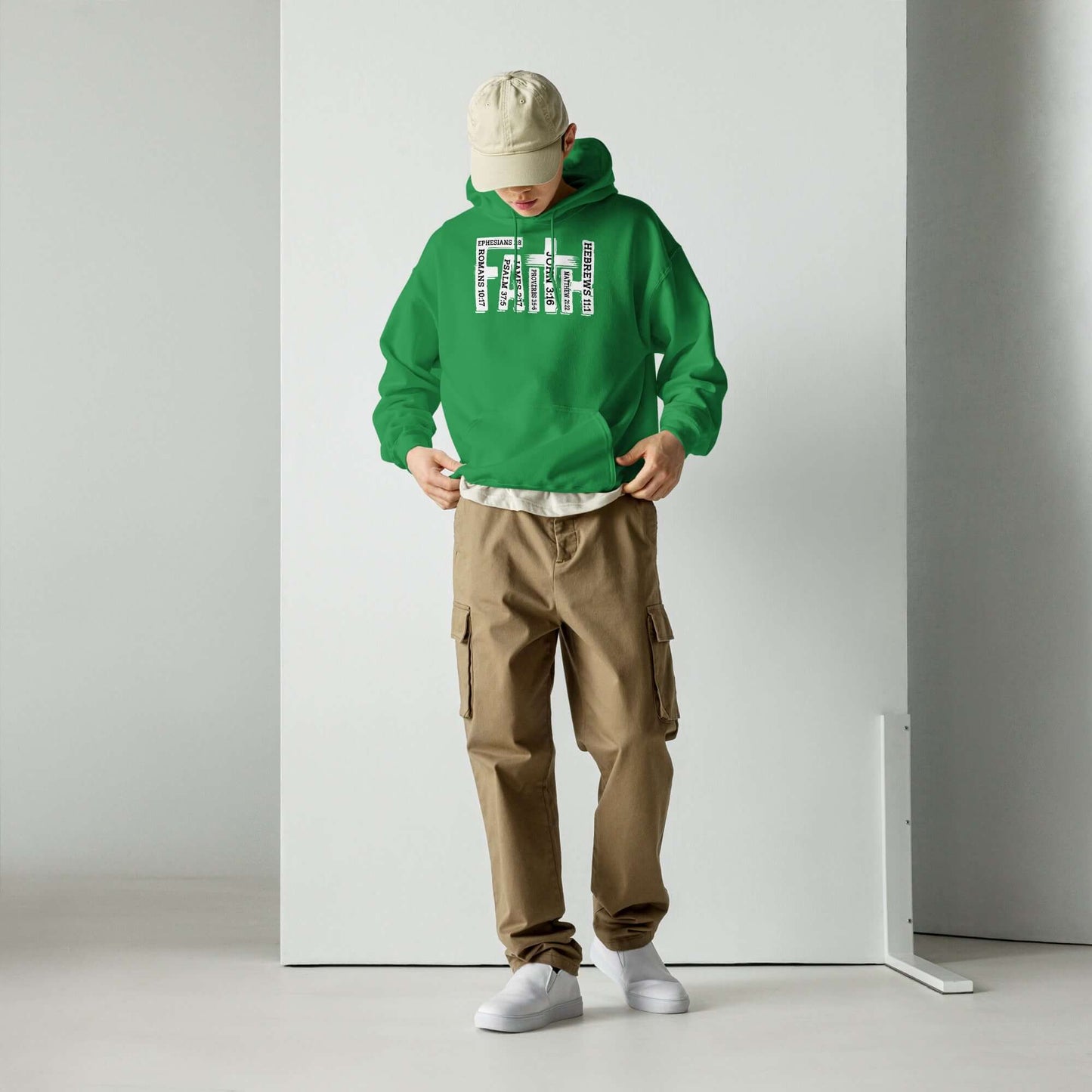 Man in green "Faith" Christian hoodie inspired by Hebrews 11:1, wearing beige cargo pants and white sneakers, looking down, standing in a minimalist setting.