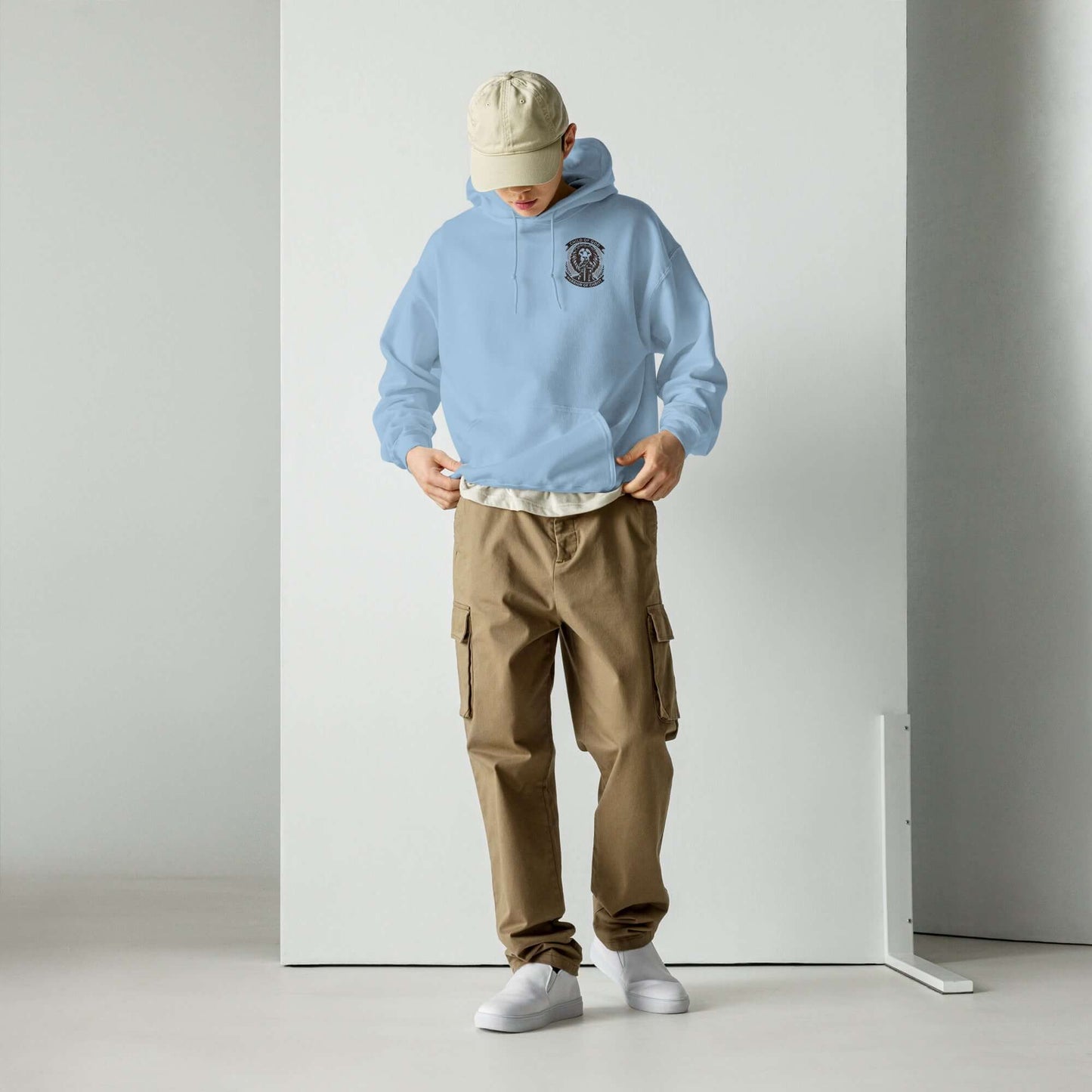 Man in blue hoodie and beige cargo pants looking down, standing in a minimalist white studio setting.