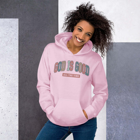 Woman wearing a pink "God is Good" hoodie, showcasing vibrant lettering and a stylish, comfortable design for Christian clothing.