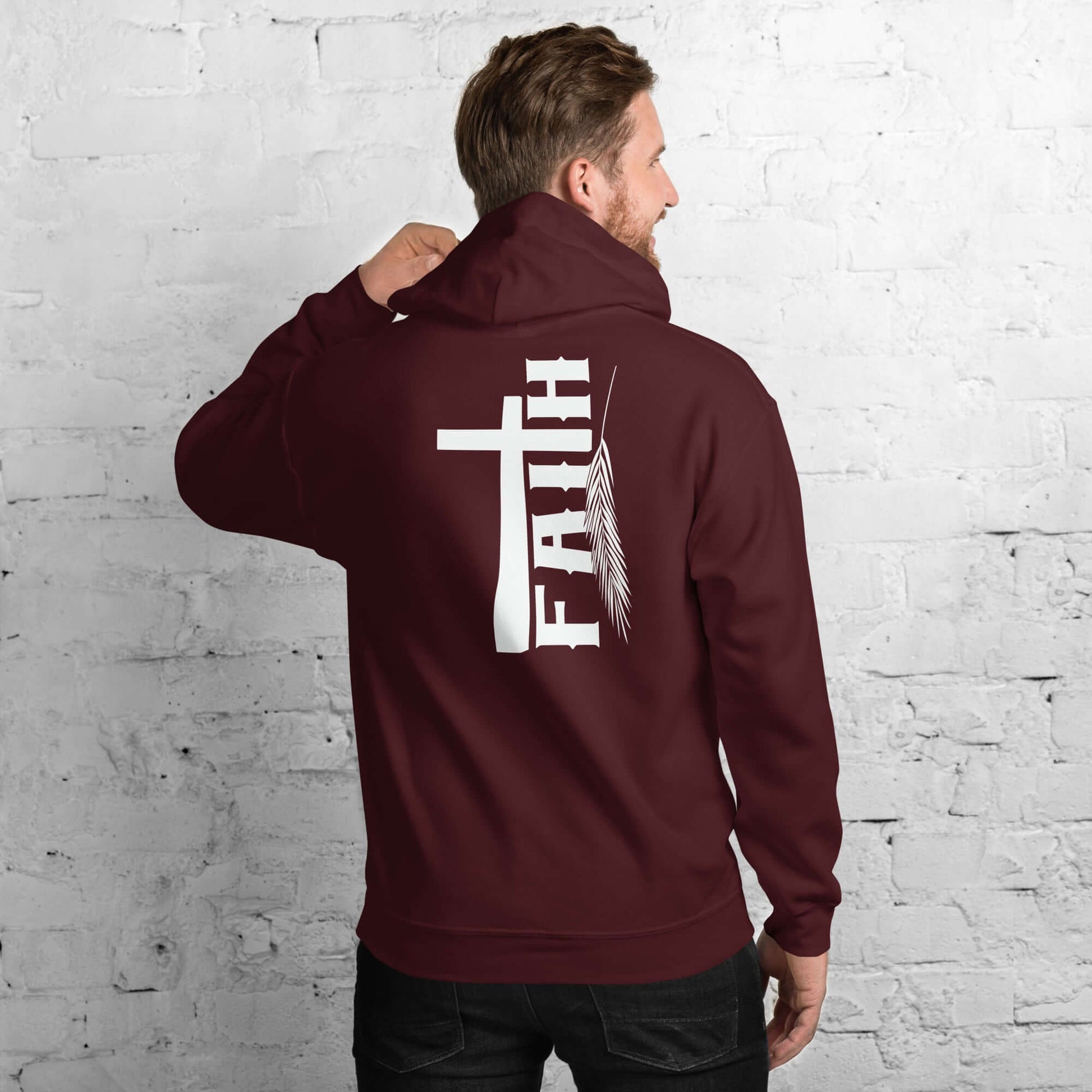 Man in maroon Faith hoodie featuring cross and feather design, inspired by Hebrews 11:1, against white brick background, Christian clothing UK.