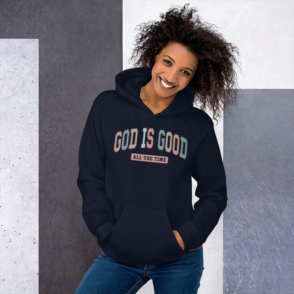 Woman wearing a dark hoodie with "God is Good" lettering, showcasing Christian clothing in a stylish and vibrant design.