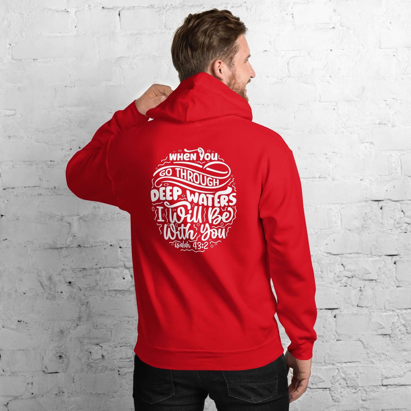 Man wearing a red "Through Deep Waters" hoodie with Isaiah 43:2 quote, Christian clothing UK, against a white brick wall.