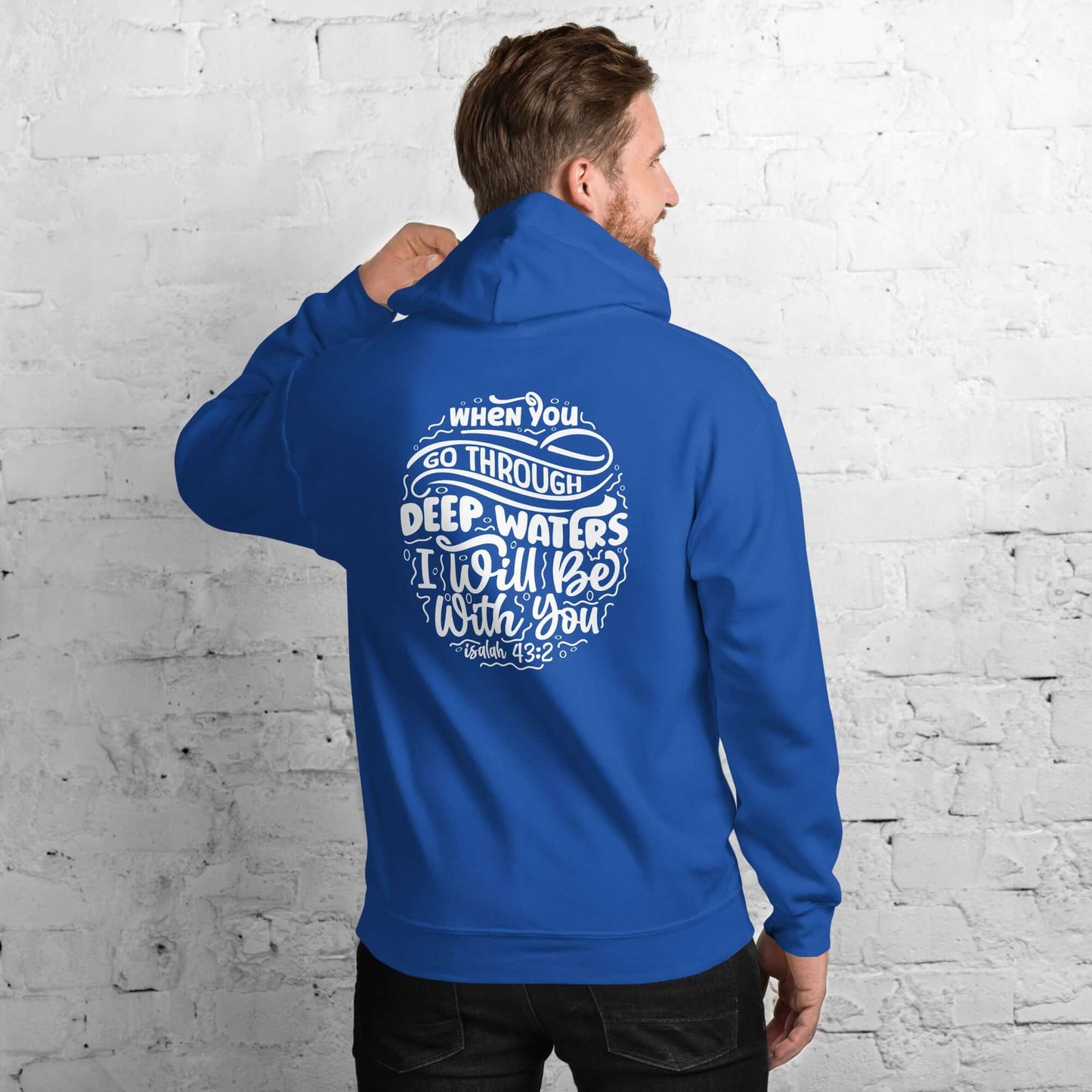 Man wearing a blue 'Through Deep Waters' hoodie with Isaiah 43:2 quote, Christian clothing UK, against a white brick background.