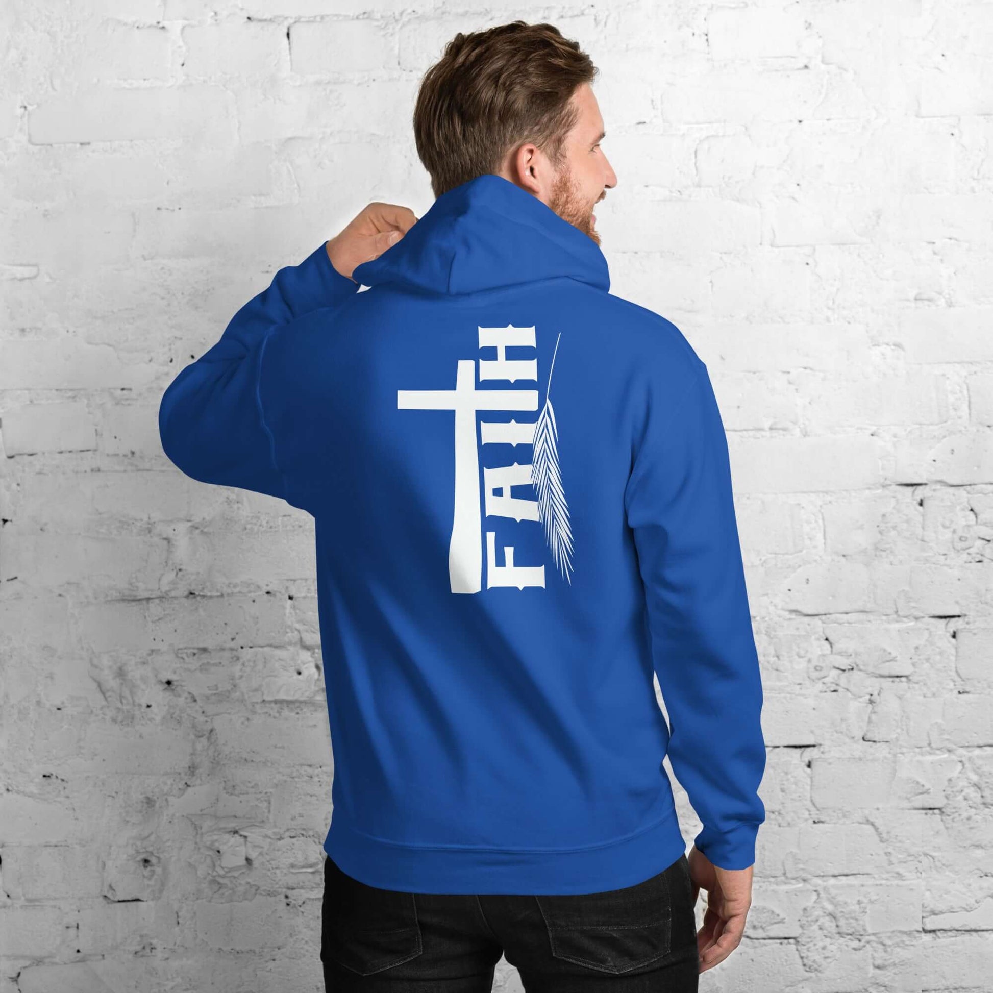 Man wearing blue 'Faith' hoodie with cross and feather design, inspired by Hebrews 11:1, against white brick background, Christian clothing UK.