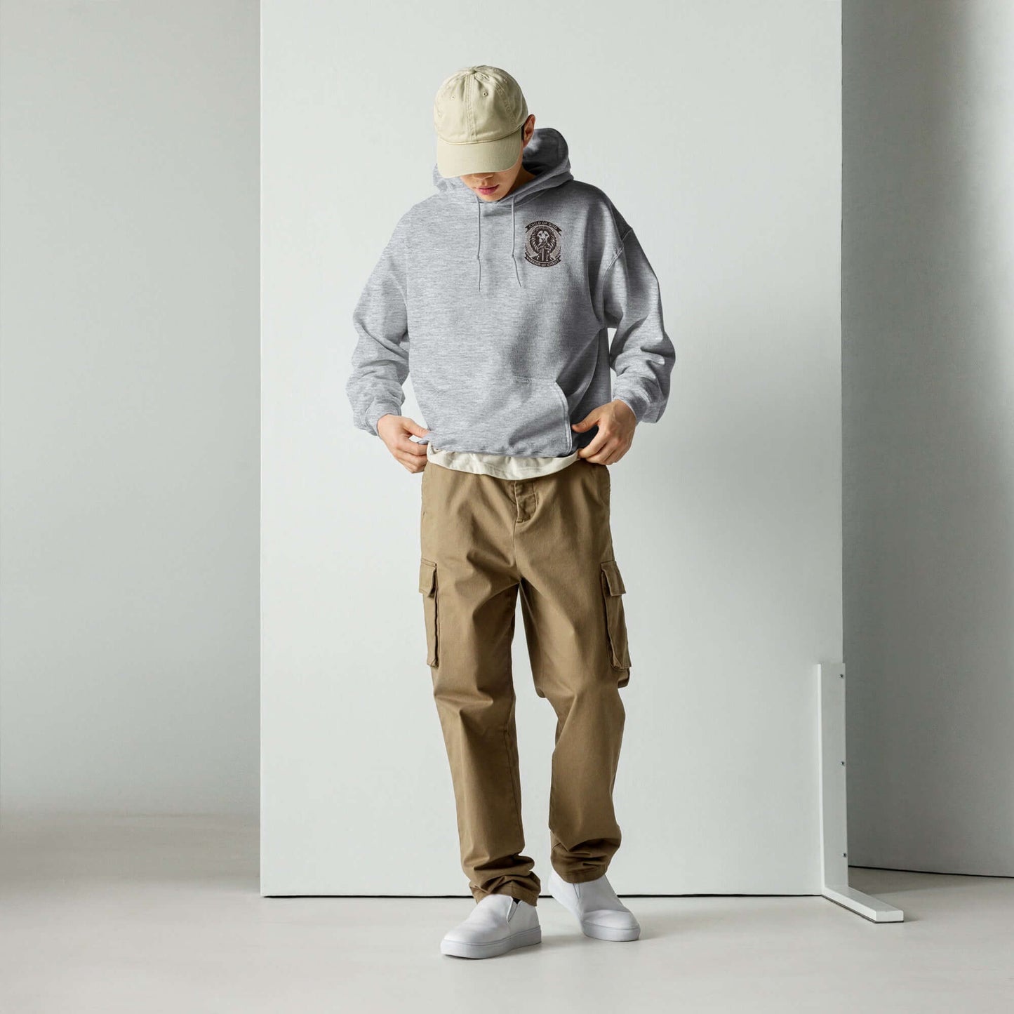 Young man in a stylish grey hoodie and beige cargo pants looking down, standing in a minimalist white space