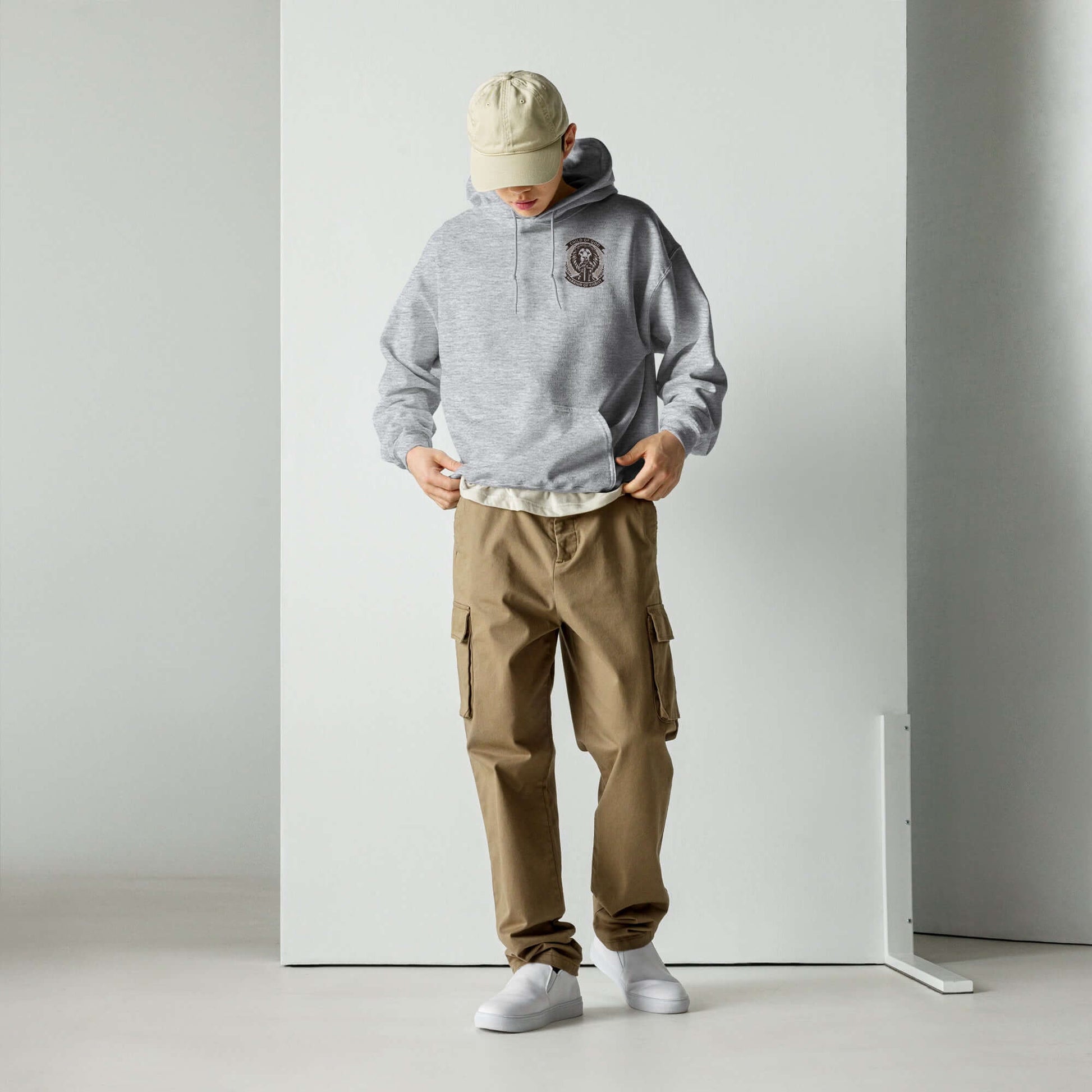 Young man in a stylish grey hoodie and beige cargo pants looking down, standing in a minimalist white space