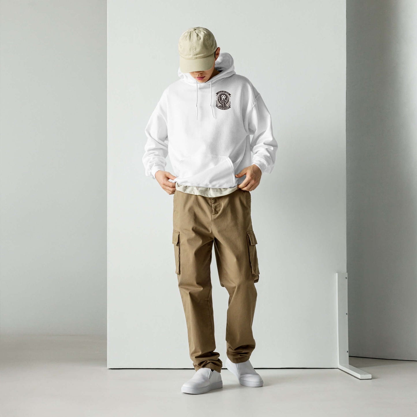 Man in white Christian hoodie and beige cargo pants looking at pockets, standing in a minimalist setting