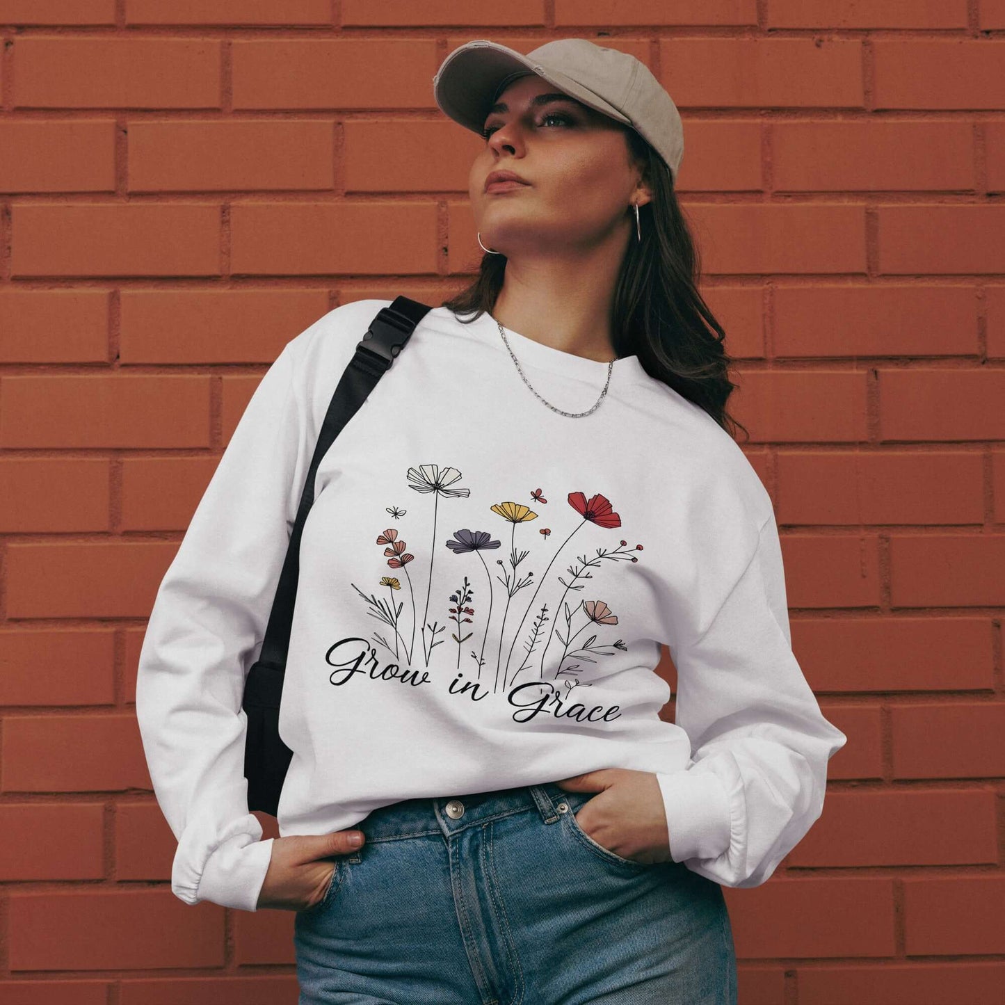 Woman wearing "Grow with Grace" Christian sweatshirt, featuring floral design, against a brick background.