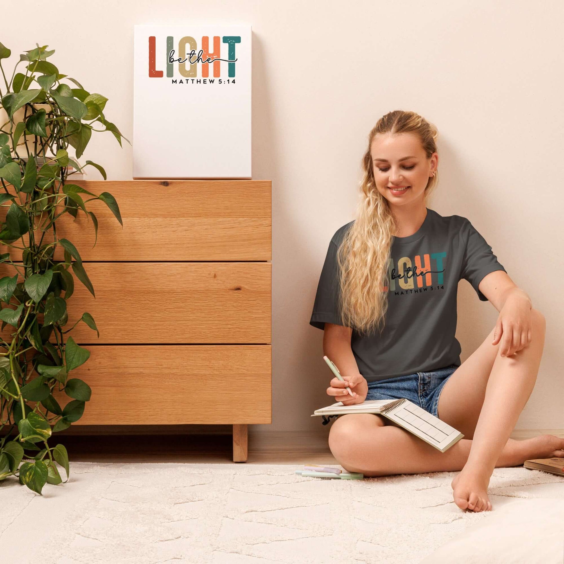 Woman wearing "Be the Light" Christian T-shirt, sitting in a cozy room with a wooden dresser and leafy plant.