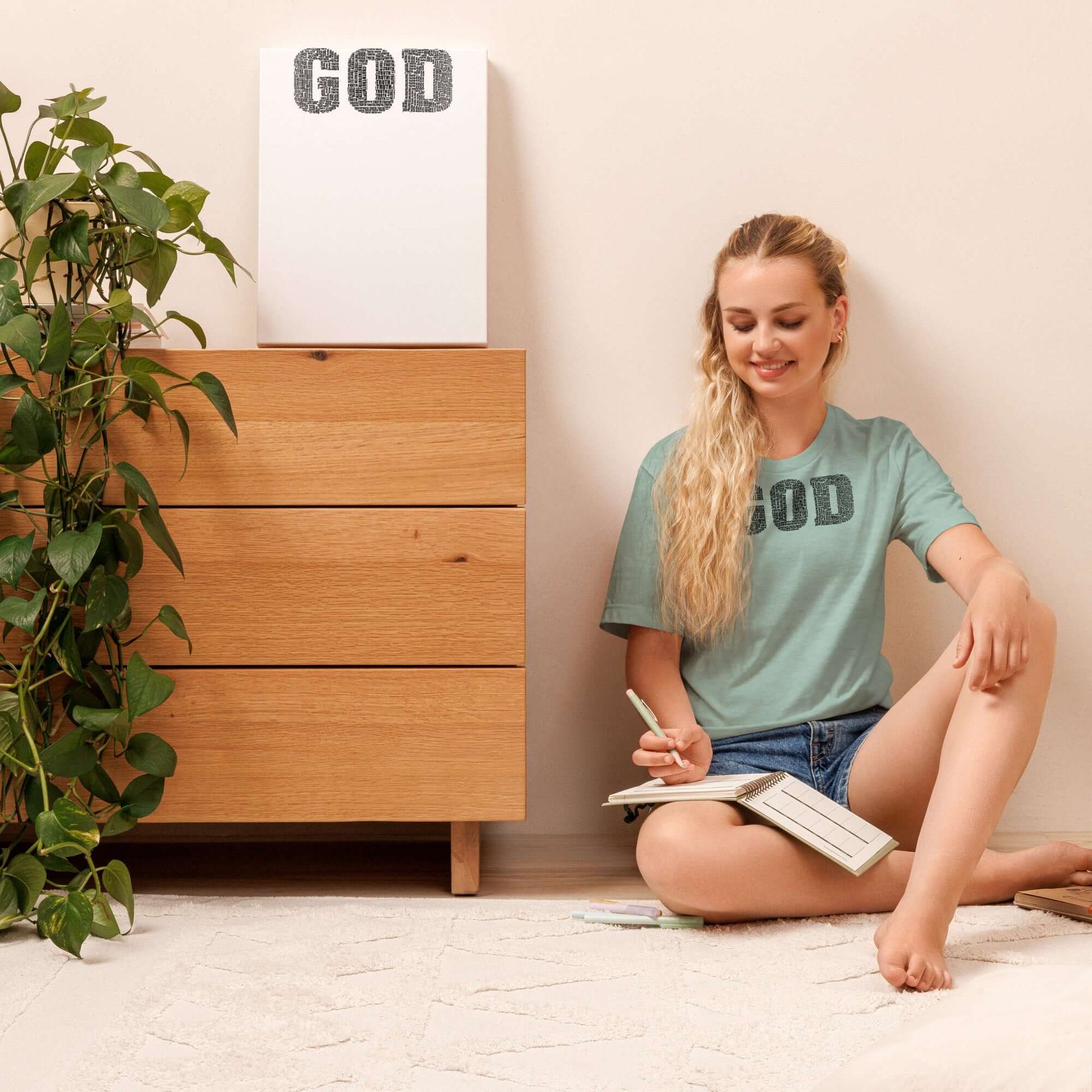 Woman in a blue "GOD" T-shirt sitting on the floor, journaling with a plant and wooden furniture, promoting Christian clothing in the UK.