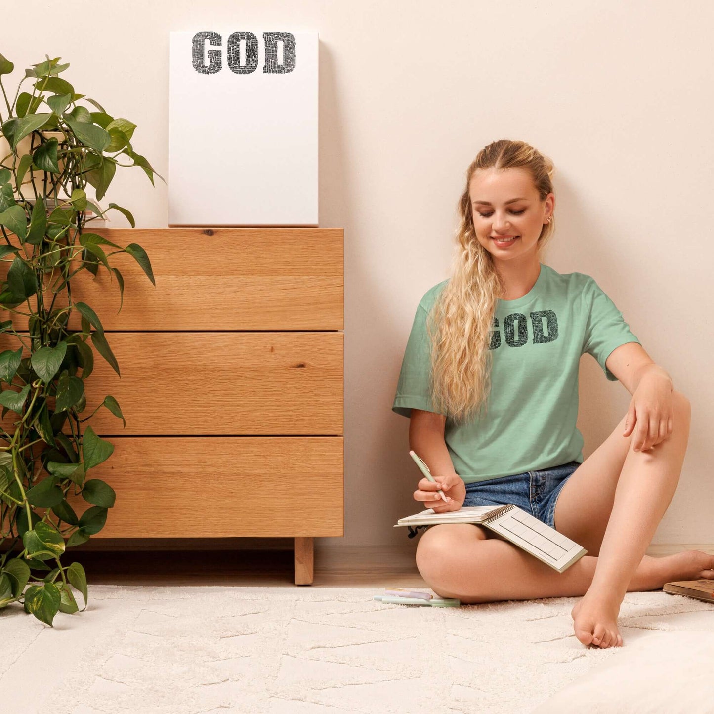 Woman wearing "GOD" Truth T-shirt, sitting on the floor with a notebook beside a wooden drawer, surrounded by greenery.