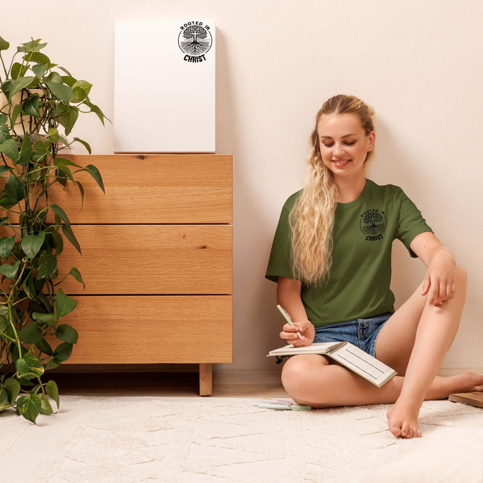 Person sitting on floor wearing Unisex Christian t-shirt "Rooted in Christ" Green, surrounded by plants and wooden furniture.