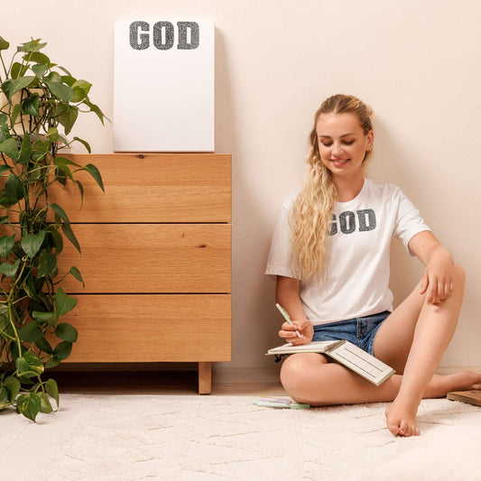 Young woman in a "GOD" Truth T-shirt sitting on the floor, writing in a notebook, showcasing Christian clothing in a cozy setting.